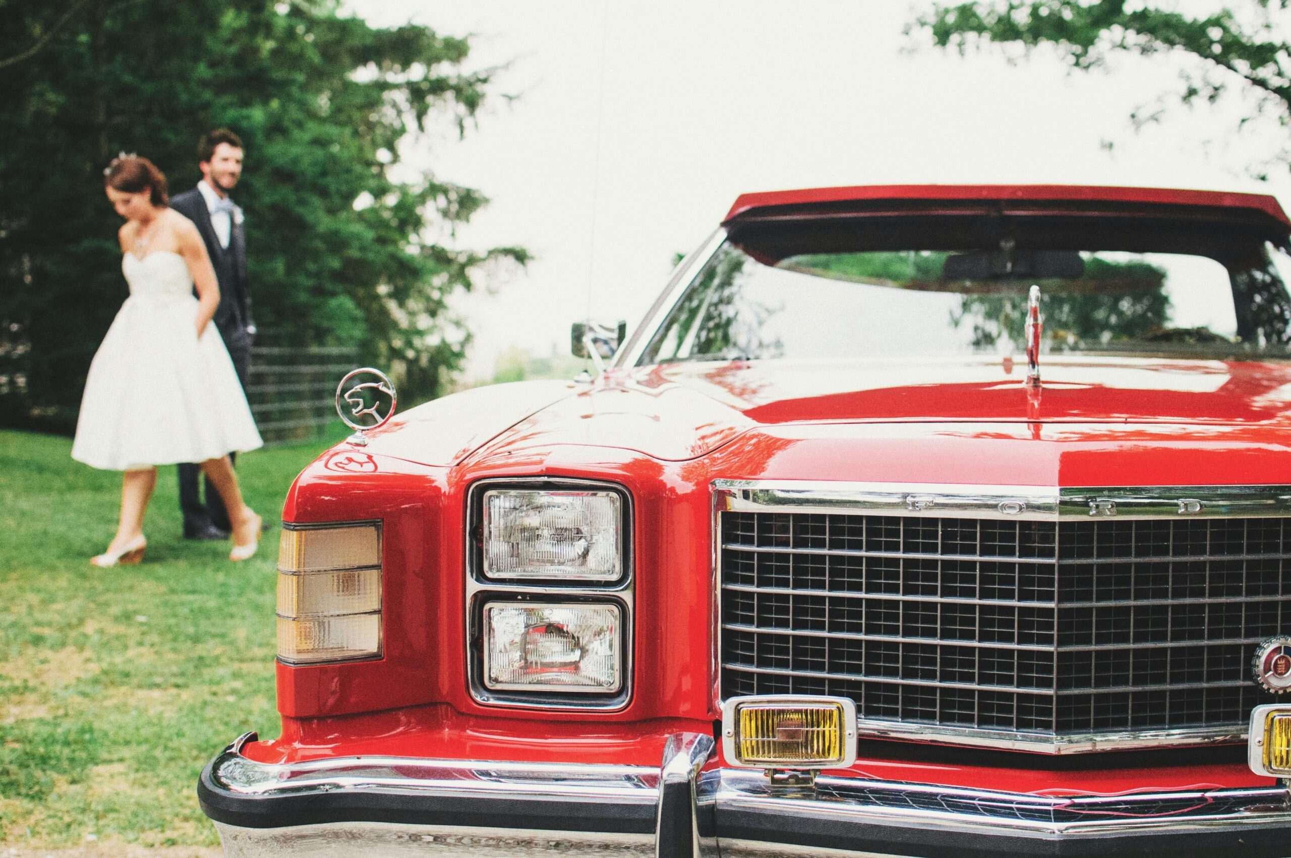 A vintage red car parked outdoors at a wedding with a bride and groom in the background.
